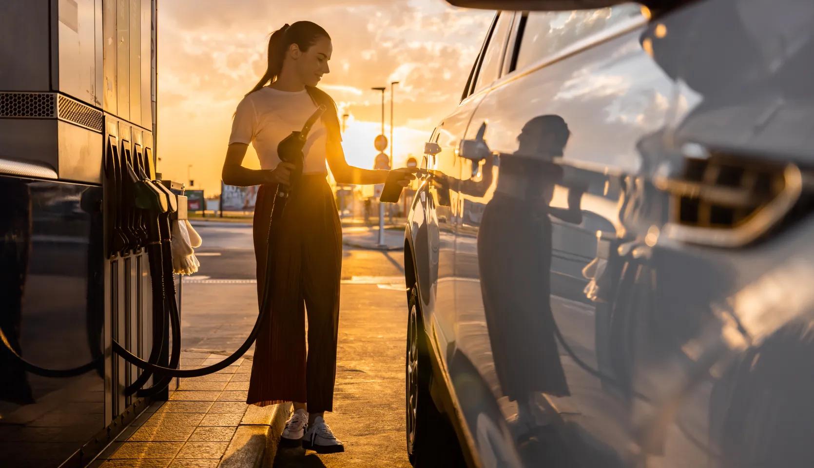 Woman fueling up her car at a gas station with a sunset in the background.
